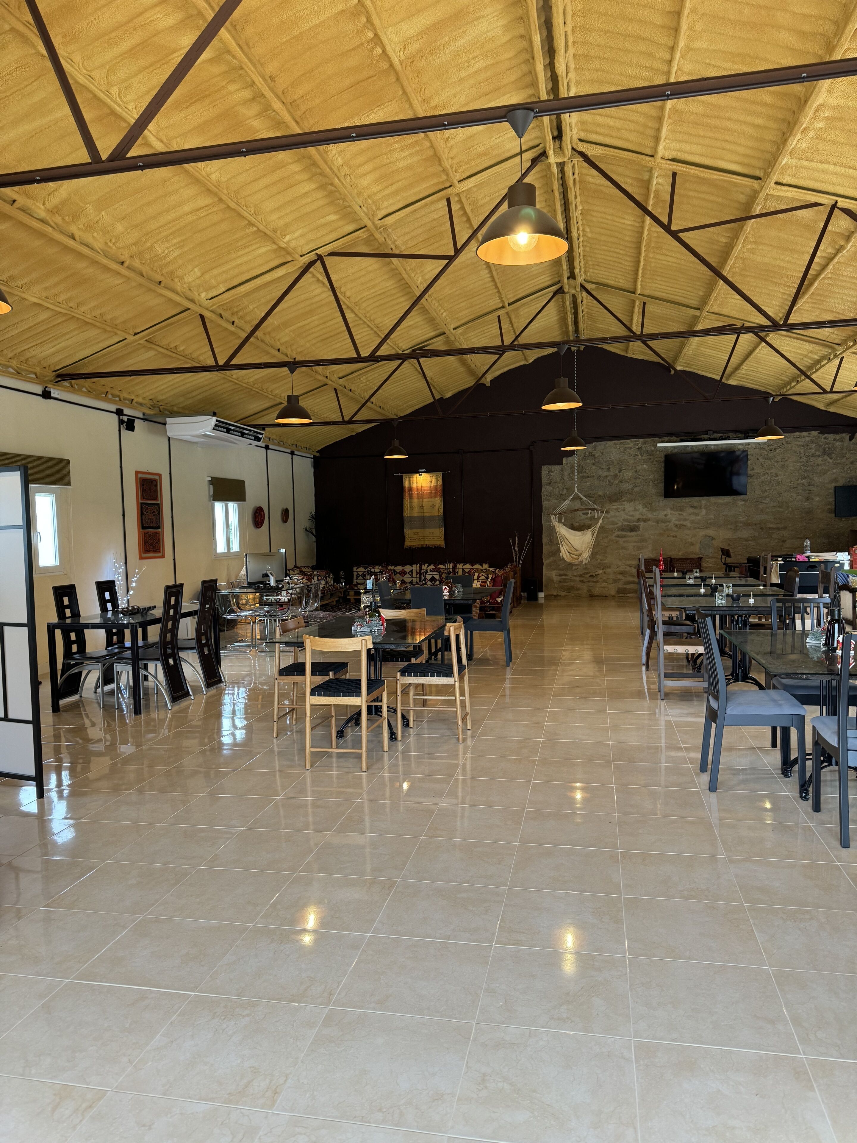 A spacious indoor area with multiple tables and chairs, a high ceiling with exposed beams, and a large screen on the wall.