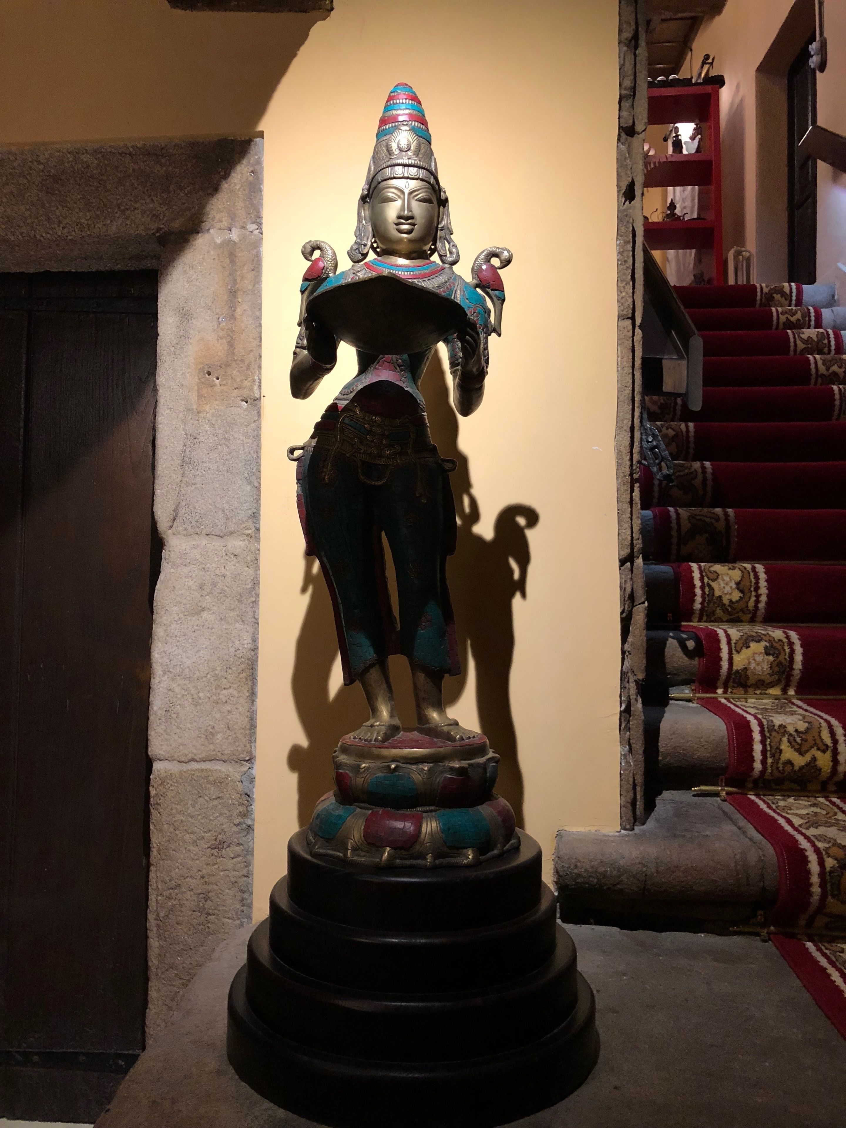 A statue of a deity with multiple arms, standing on a multi-tiered base, positioned against a wall with a staircase in the background.