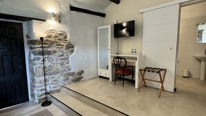 Una habitación con pared de piedra, una puerta negra, una puerta blanca, un televisor montado en la pared, una silla, una mesita y un banco.