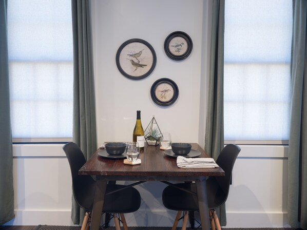 Une table à manger avec des chaises, une bouteille de vin, des bols et un verre. Il y a trois tableaux encadrés accrochés au mur.
