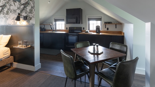 Un dormitorio estilo loft acogedor con cama, mesita de noche y velador. La habitación tiene piso de madera oscura, pared con diseño y ventana con persianas.
