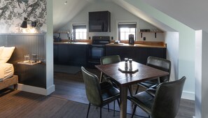 Une chambre à coucher chaleureuse, aménagée dans un style loft, avec un lit, une table de chevet et une commode. La pièce est dotée d’un plancher en bois foncé, d’un mur à motifs et d’une fenêtre avec des stores.
