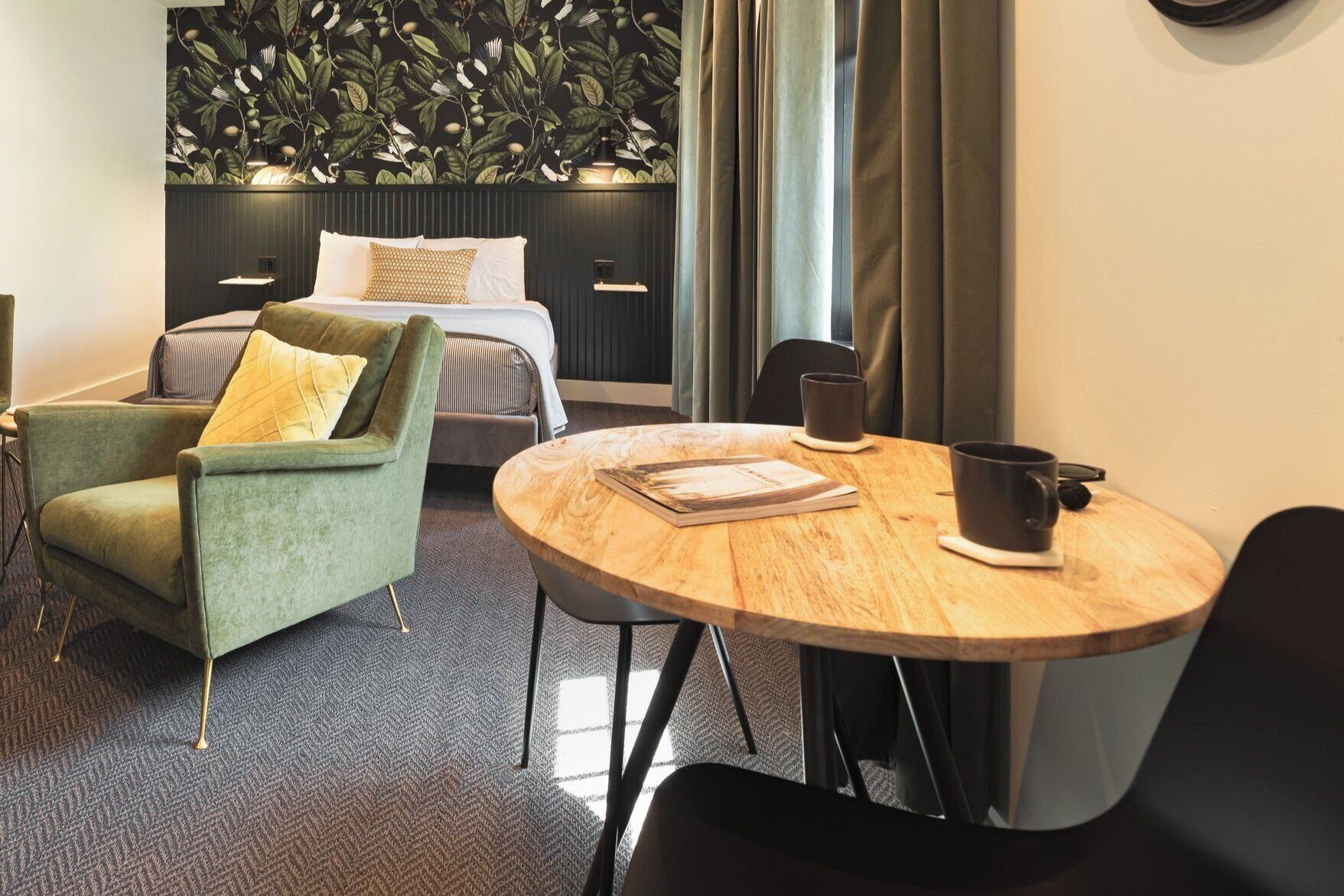 Quarto de hotel com uma cama, uma poltrona verde, uma mesa redonda de madeira e uma janela grande com cortinas.