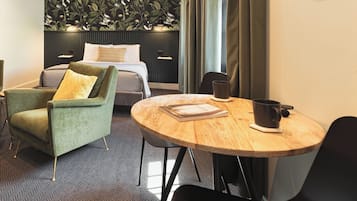 Quarto de hotel com uma cama, uma poltrona verde, uma mesa redonda de madeira e uma janela grande com cortinas.