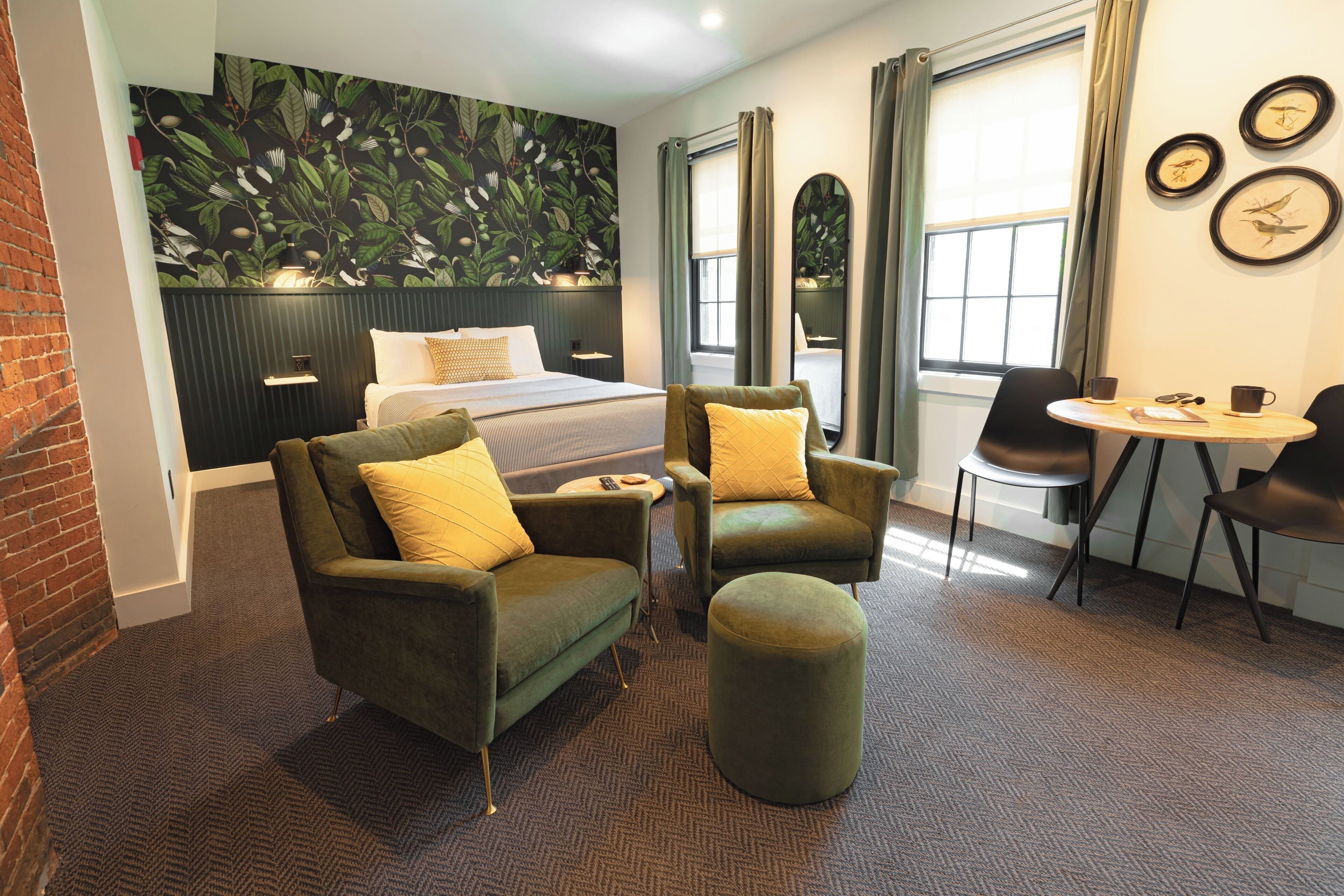 Habitación de hotel con cama, dos sillones, un taburete redondo, una mesita y una ventana con cortinas.