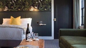 Una habitación de hotel con una cama, una mesa de centro de madera y un posavasos de mármol, una botella y vasos, un sofá verde y una pared con un estampado de hojas.
