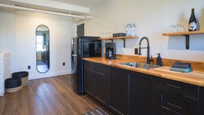 Una cocina moderna con armarios oscuros, encimera de madera, pileta integrada y cafetera. Hay estantes con botellas y vasos, y un espejo en la pared.