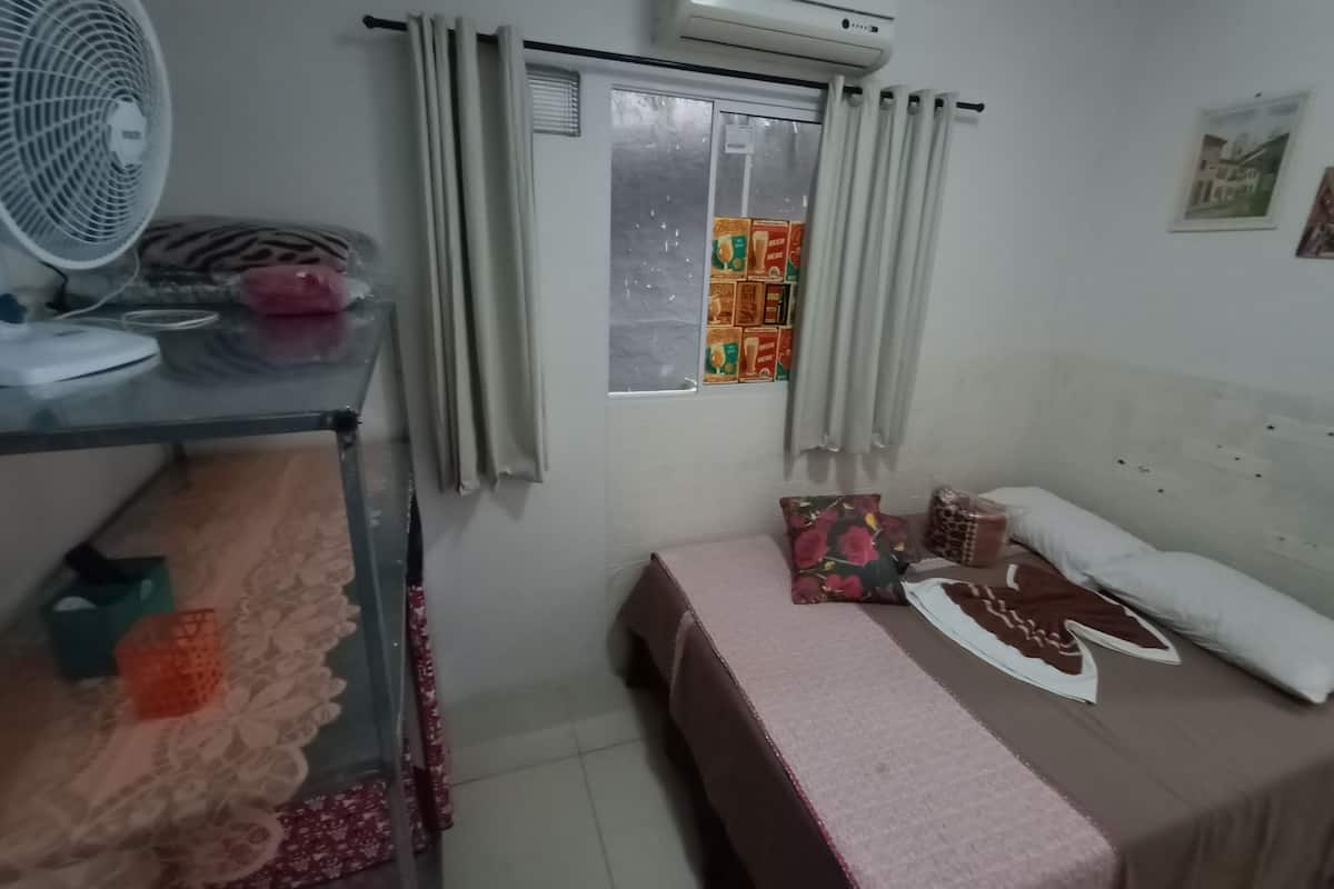 A bedroom with a bed, a bedside table, a fan, a window with curtains, and a wall-mounted air conditioner.