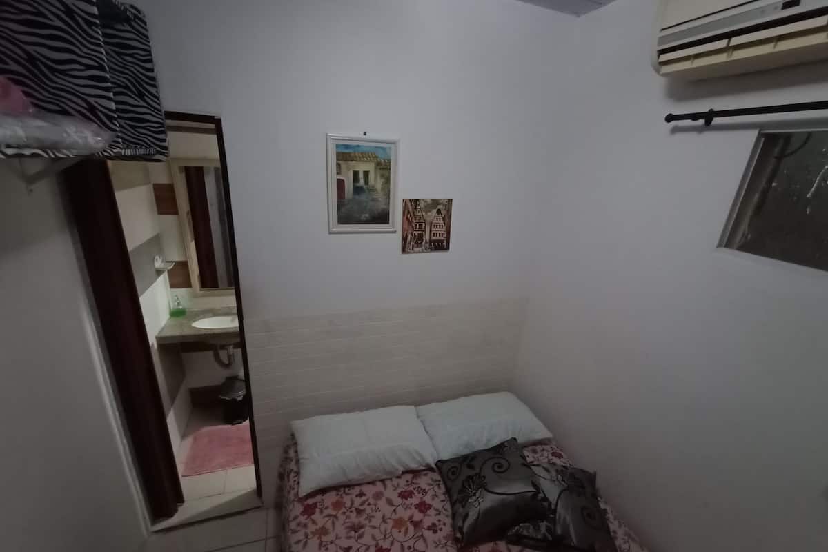 A small, dimly lit room with a bed, a mirror, and a window.