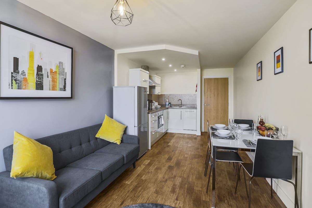 A modern apartment with a grey sofa, yellow cushions, a dining area with a table set for four, and a kitchen in the background.