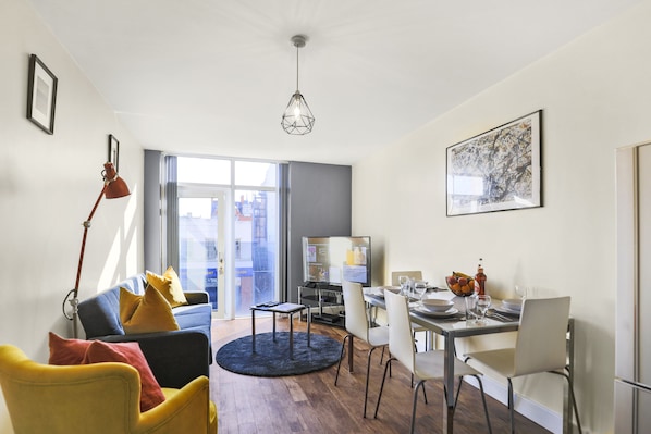 A modern living room with a sofa, dining table, and chairs. There is a television and a window with a view of buildings.