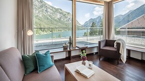 A modern room with a large window overlooking a lake and mountains, wooden furniture, and a cozy seating area. - Hotel Fischer am See (Heiterwang)