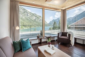 Una habitación moderna con un gran ventanal con vistas a un lago y montañas, muebles de madera y una zona de estar acogedora.