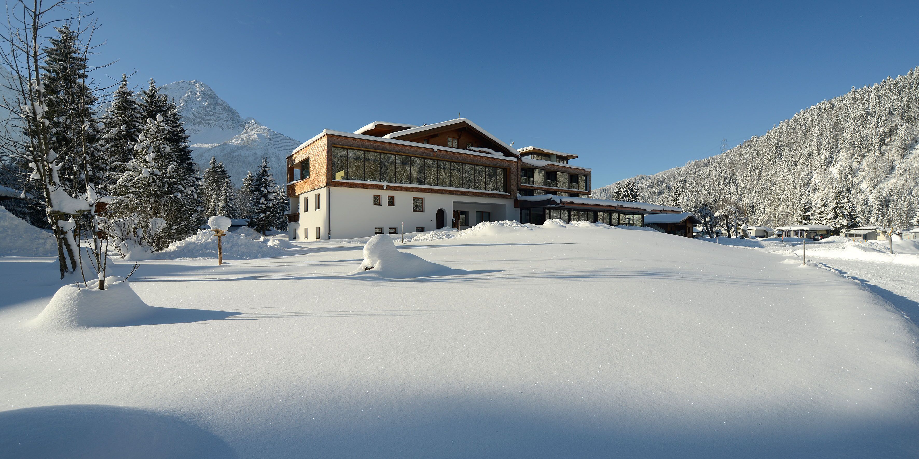 Un moderno edificio de estación de esquí con grandes ventanales, rodeado de árboles nevados y montañas.