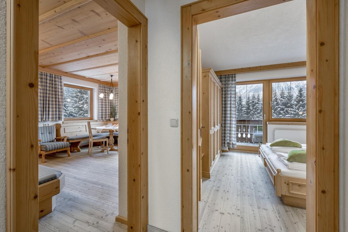 A wooden interior with a dining area, a sofa, and a bed with a view of snow-covered trees.