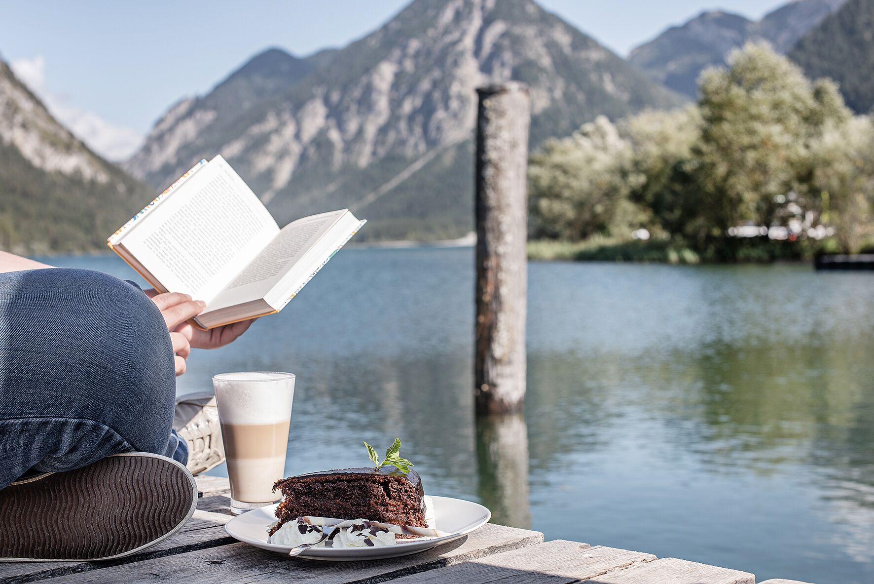 Una persona leyendo un libro a orillas de un lago, con una rebanada de pastel y una taza de café sobre una mesa de madera.