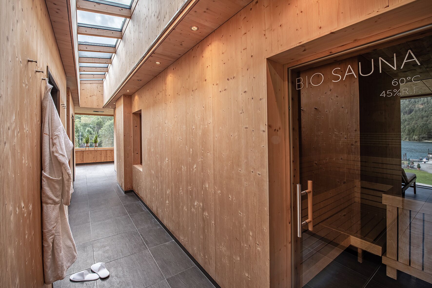 Un couloir en bois mène à un sauna doté d’une porte en verre, une serviette est accrochée au mur et une paire de chaussons est posée sur le sol.