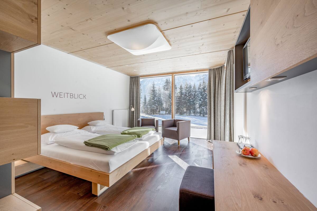 Moderni hotellihuone, jossa on puinen katto, suuri sänky valkoisilla vuodevaatteilla, vihreä peitto, puinen työpöytä, jossa on lautanen täynnä hedelmiä, ja suuret ikkunat, joista avautuu näkymä lumipeitteisiin puihin.