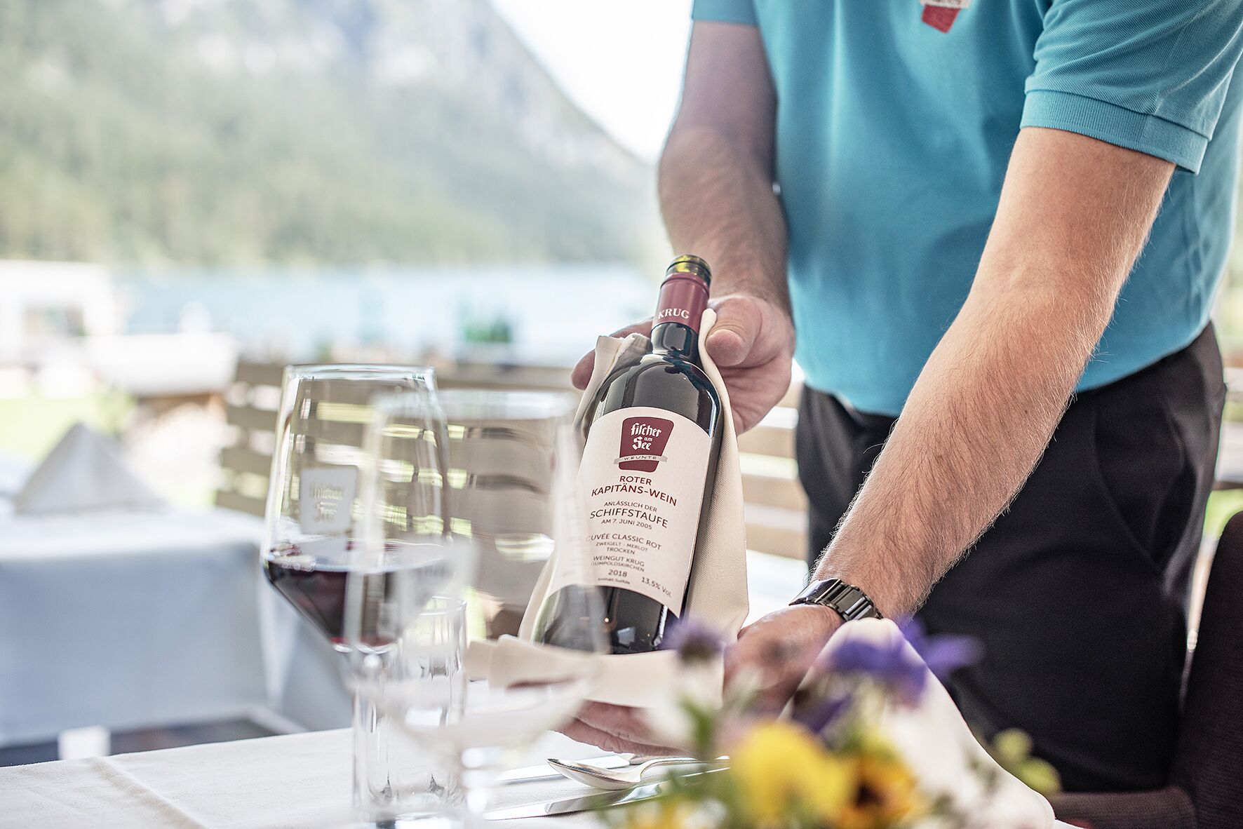 Eine Person gießt Wein in ein Glas an einem Tisch, im Hintergrund eine Berglandschaft.