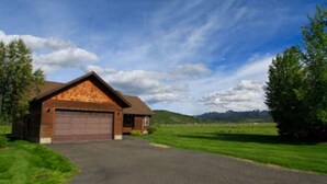 Ein eingeschossiges Holzhaus mit Garage, umgeben von gepflegtem Rasen und Bäumen, vor einer Hügellandschaft und einem teils bewölkten Himmel.