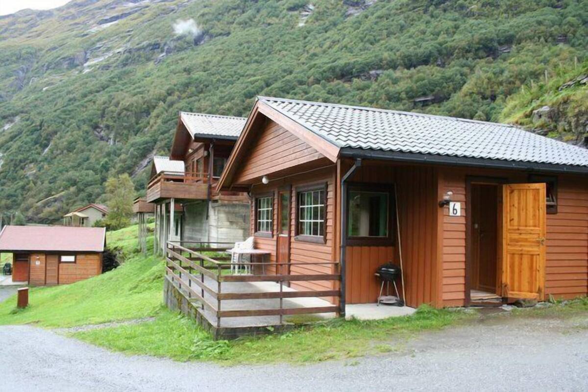 A wooden cabin with a porch and a small table, surrounded by lush greenery and mountains.