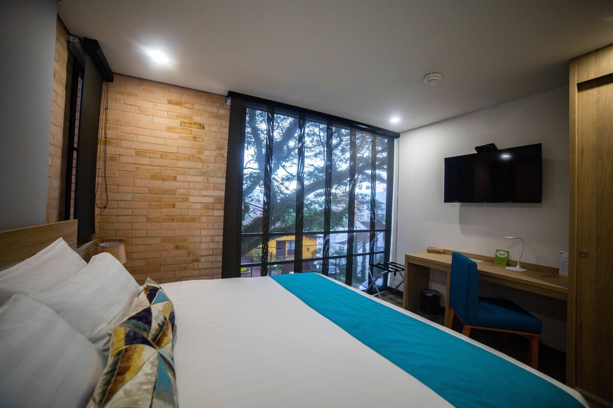 Une chambre d’hôtel équipée d’un lit, d’un bureau, d’une chaise, d’une télévision et d’une fenêtre donnant sur des arbres.