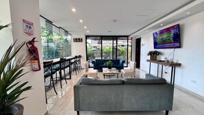 A modern living room with a grey sofa, a coffee table, and a TV mounted on the wall. There is a fire extinguisher mounted on the wall and a plant on the left side. - EcoHub Hotel Medellin (Medellín)