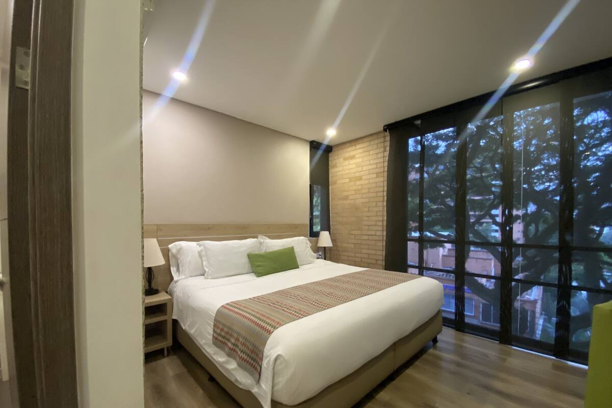 Une chambre d’hôtel moderne avec un grand lit, un plancher en bois, un mur décoratif en briques et une vue sur les arbres par une grande fenêtre.