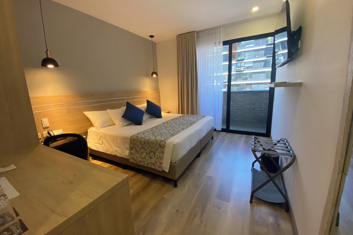 Habitación de hotel con cama, mesita de noche, silla y vistas al exterior.