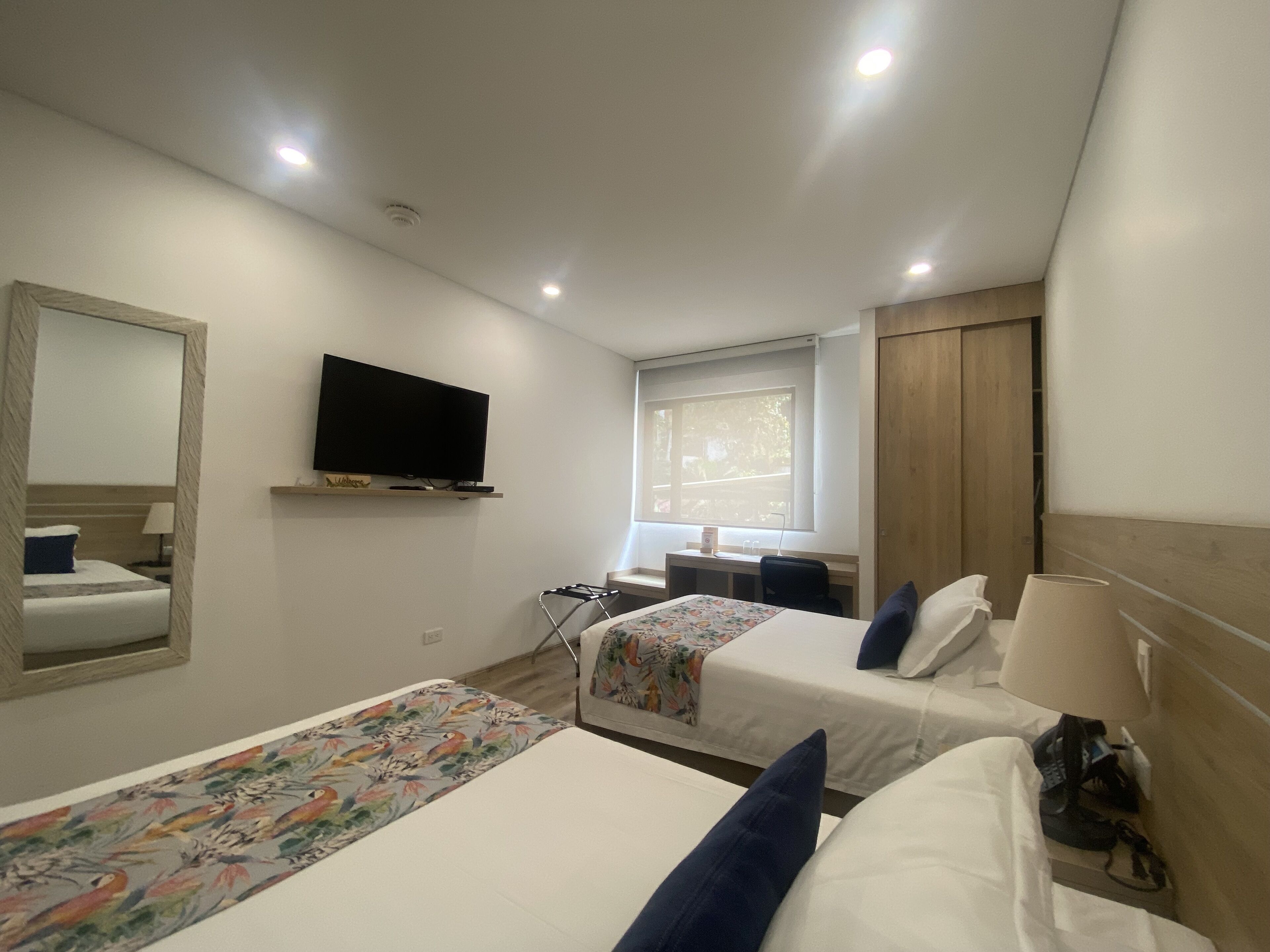 A hotel room with two beds, a TV mounted on the wall, a desk with a chair, and a window providing natural light.