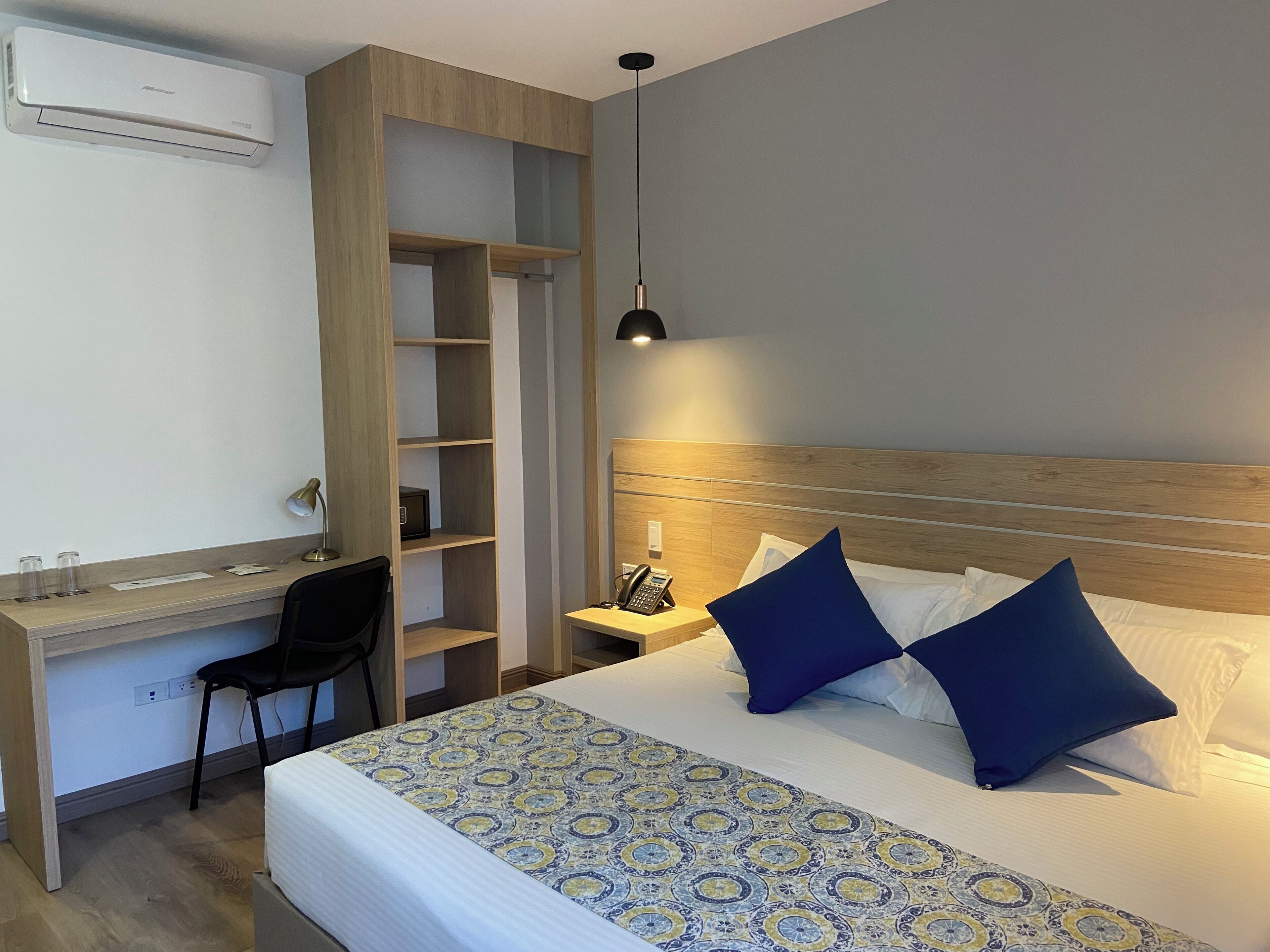 Una habitación de hotel con cama, mesita de noche, lámpara, escritorio, silla y armario empotrado.