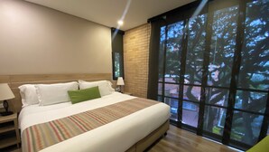 A modern hotel room with a large bed, a nightstand, a lamp, and a view of trees through the window. - EcoHub Hotel Medellin (Medellín)
