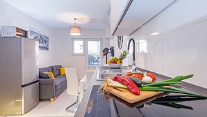 A modern kitchen with a built-in oven, a dining area with a grey sofa, and a view of the city through the window. - Apartment Sendi - Near the beach, fully equipped (Dubrovnik)