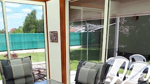 Een terras buiten, met een tafel en stoelen met een glazen blad, omgeven door een houten terras en een schuifpui die uitkomt op een achtertuin.