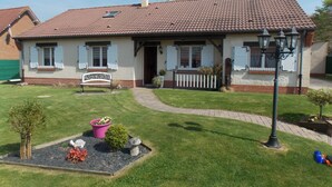 Een huis met twee verdiepingen, voorzien van een pannendak, een voortuin met een bankje en een kleine tuin met een pad dat naar de ingang leidt.