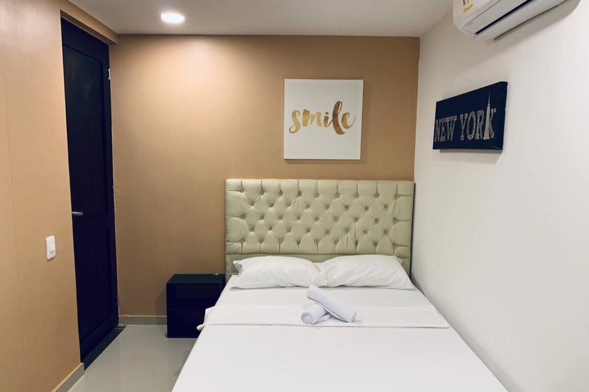 A hotel room with a bed, a headboard, a wall with a 'smile' sign, and a 'NEW YORK' sign.