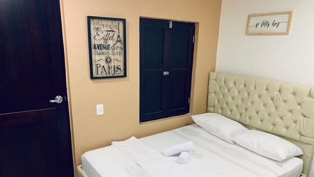 Un lit bien fait, avec du linge de lit blanc et une serviette enroulée, une tête de lit capitonnée, une porte sombre et un mur orné d’un tableau.