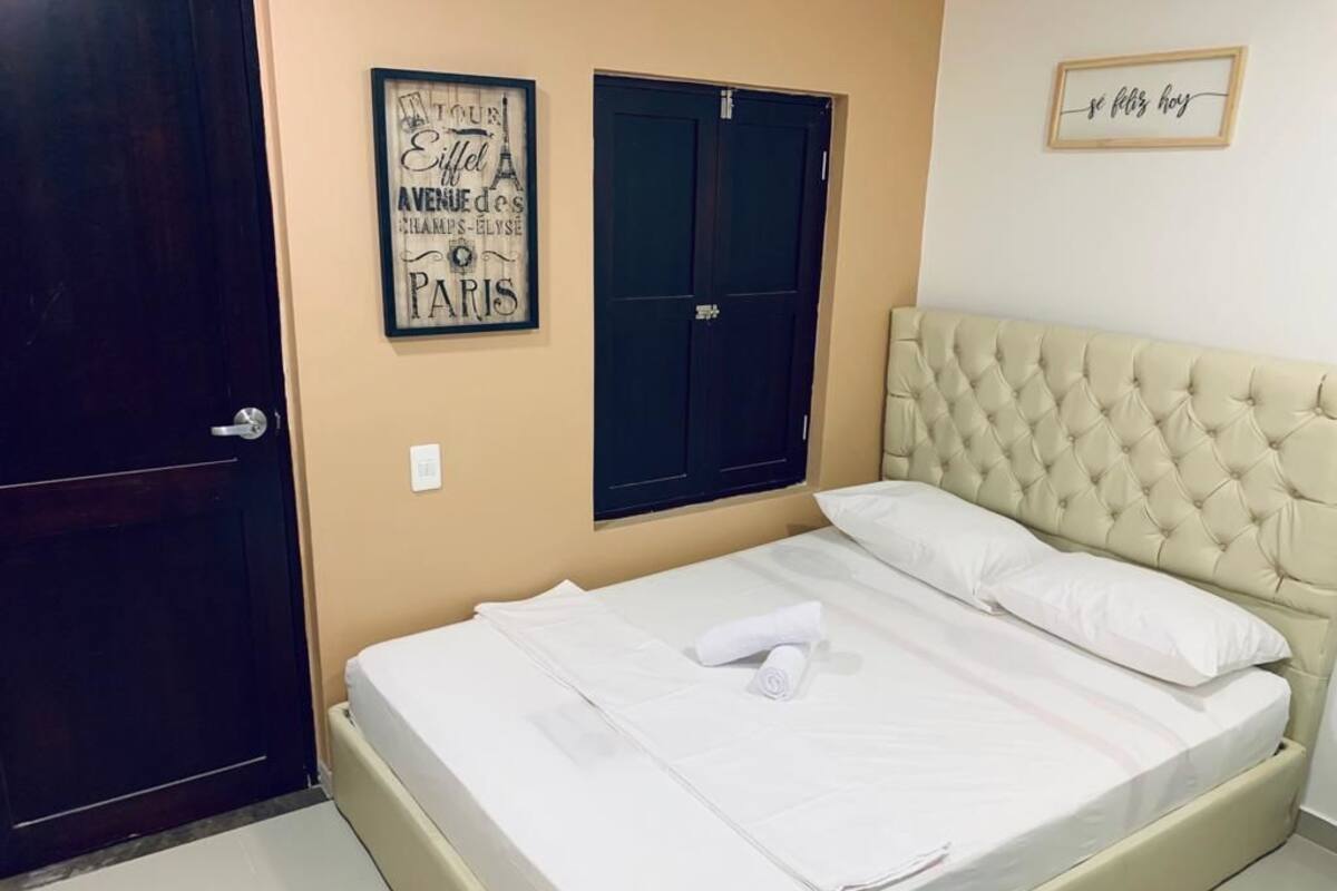 A neatly made bed with white linens and a rolled towel, a tufted headboard, a dark door, and a wall with a framed sign.