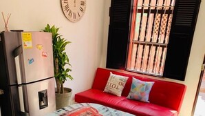 Un espace de vie compact comprenant une table à manger, des chaises, un canapé rouge, une horloge, un réfrigérateur et une fenêtre avec des barreaux.