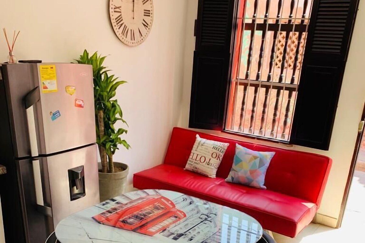 A compact living space with a dining table, chairs, a red sofa, a clock, a refrigerator, and a window with bars.