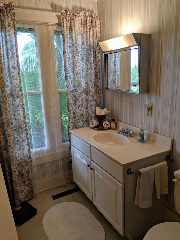 Ein Badezimmer mit einem weißen Waschbecken, einem Spiegel darüber und einer Toilette. Ein Fenster mit floralem Vorhang und ein weißer Teppich sind ebenfalls vorhanden.
