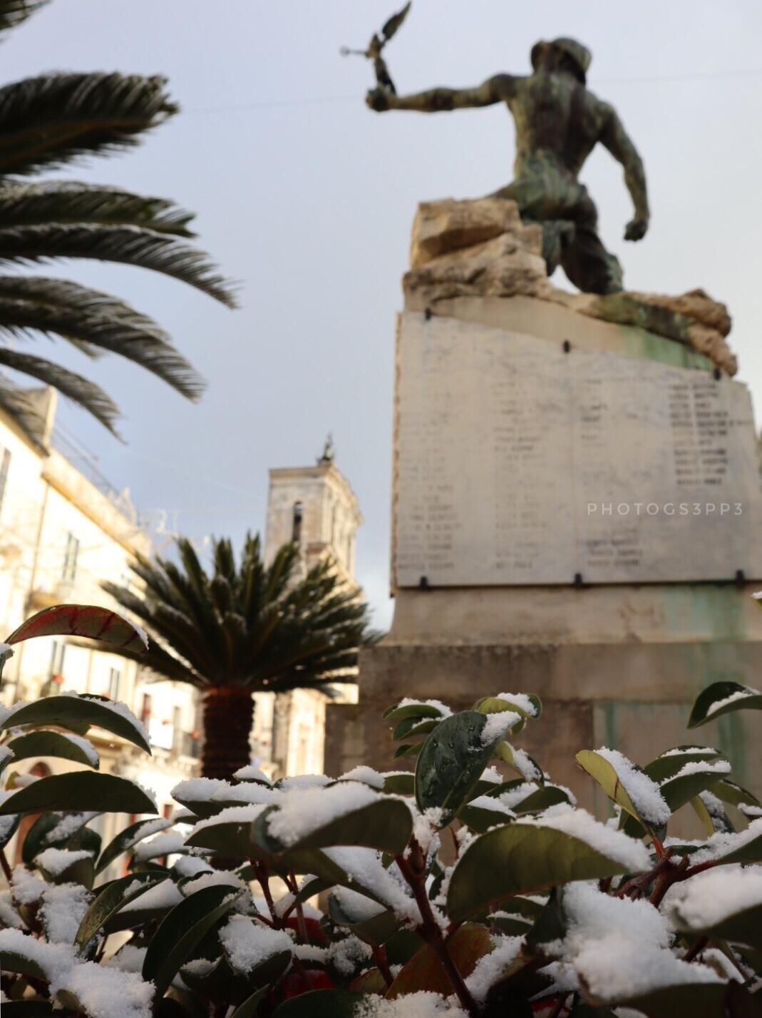 A statue of a figure with a bird, surrounded by snow-covered plants, with a building and a tower in the background.