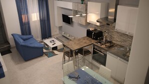 Una cocina moderna con estufa integrada, zona de comedor con mesa y sillas, un sofá azul y un televisor montado en la pared.