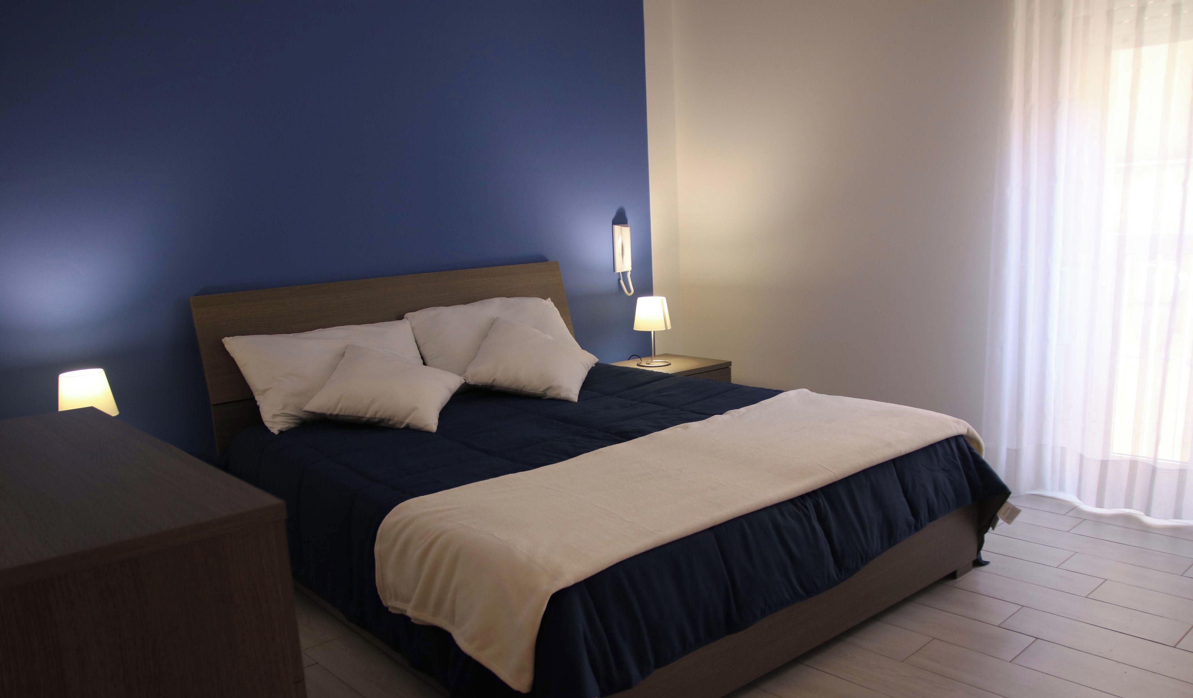 A neatly made bed with a dark blue headboard, a beige blanket, and white pillows. There are two bedside tables with lamps, one on each side of the bed. The room has light wooden flooring and a window with sheer curtains.