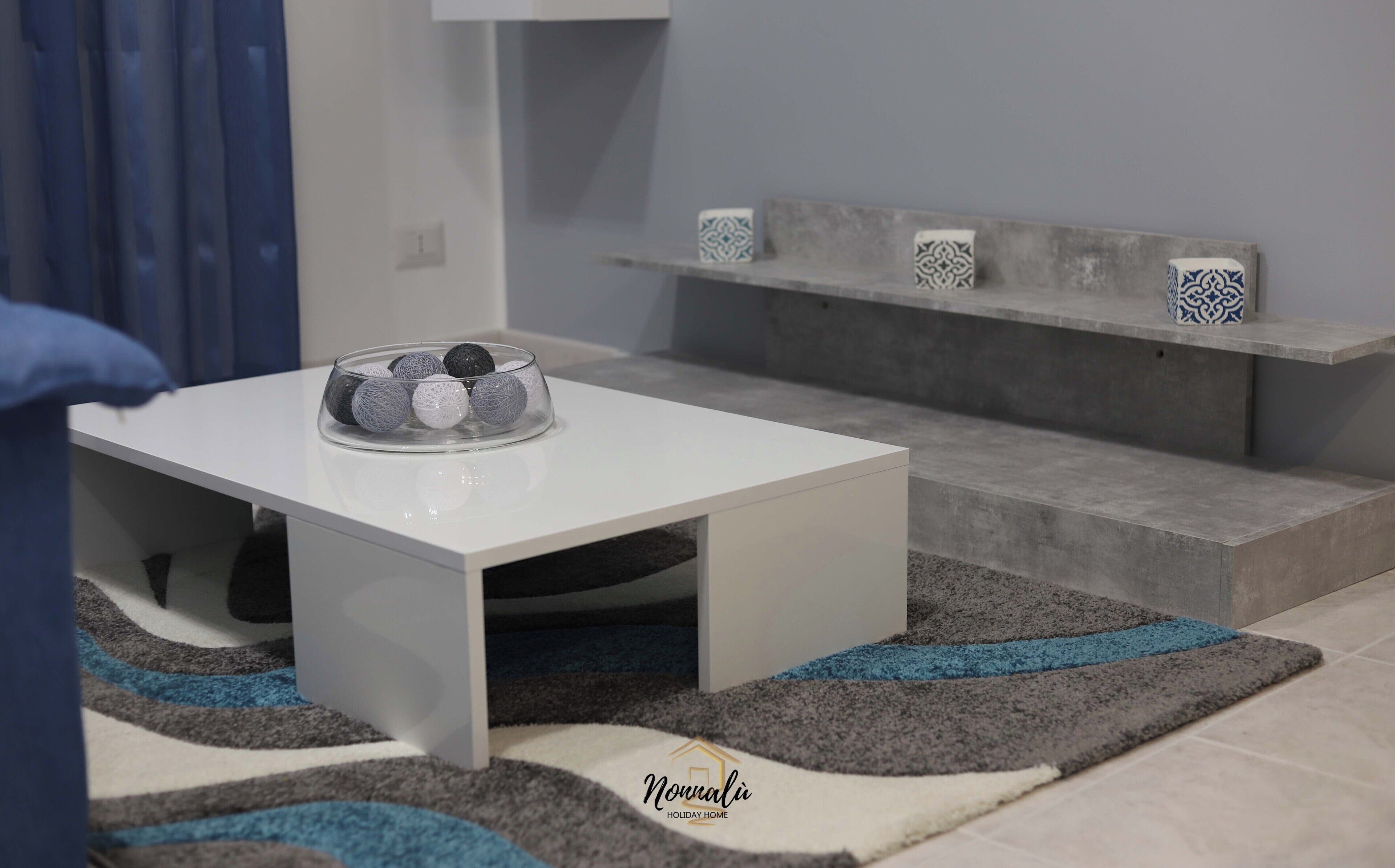 A modern living room with a white coffee table, a grey sofa, and a patterned rug.