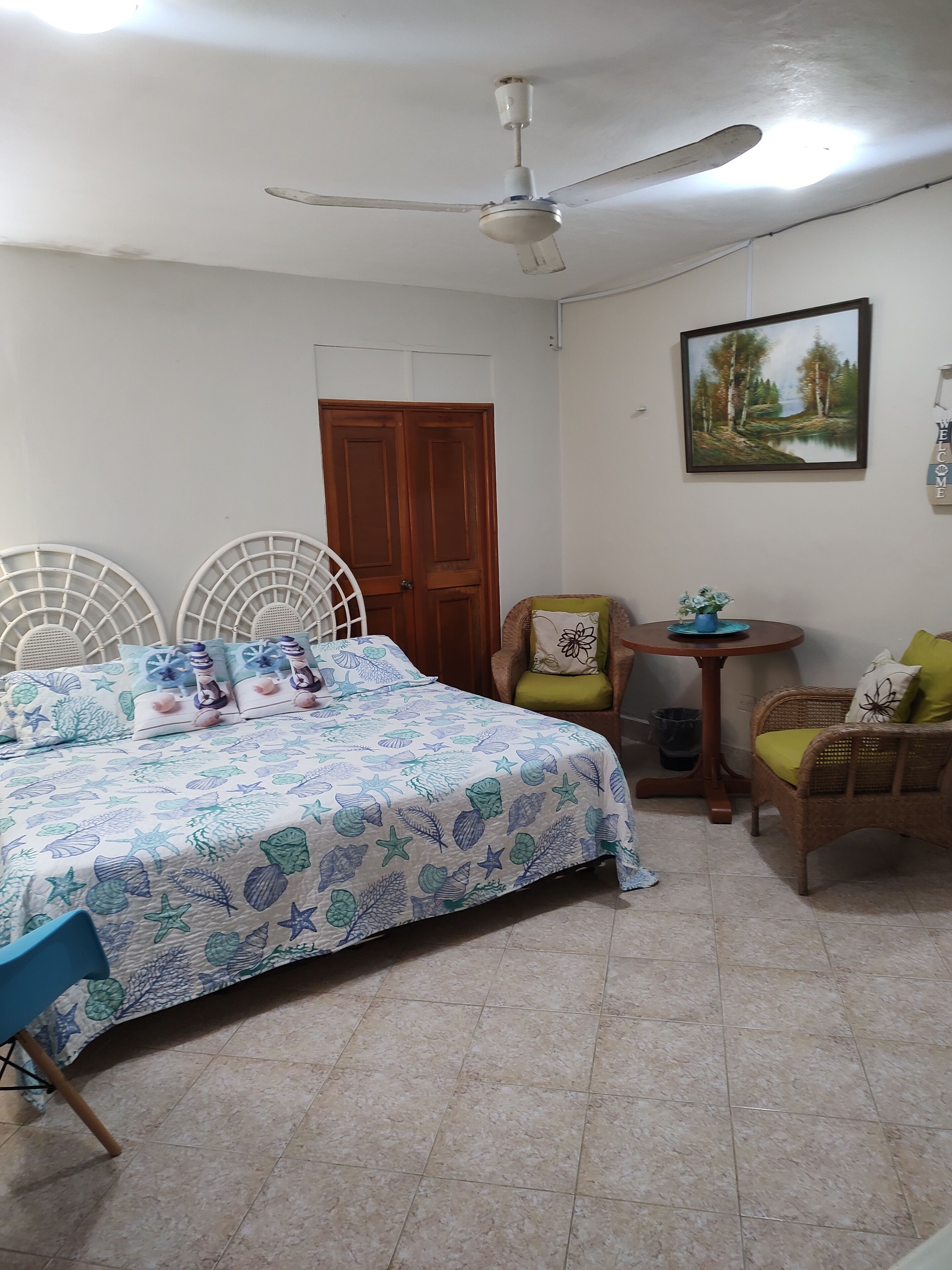 A bedroom with a bed, two chairs, a small table, a ceiling fan, a door, and a painting on the wall.
