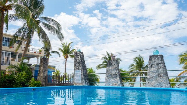 A swimming pool with a clear view of the ocean, surrounded by palm trees and a building with balconies. - Cozy Studio/Room Enclosed on an Oceanfront Property at Mima´s Village Cozumel (San Miguel de Cozumel)
