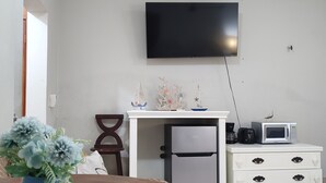 A compact living space with a wooden dining table, a white dresser, a small refrigerator, a microwave, and a flat-screen TV mounted on the wall. - Cozy Studio/Room Enclosed on an Oceanfront Property at Mima´s Village Cozumel (San Miguel de Cozumel)