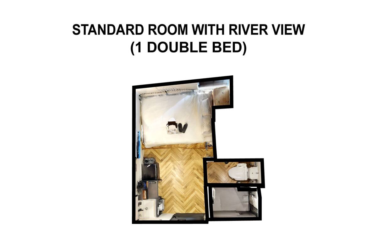 A standard room with a double bed, a wooden floor, a small table, a chair, a bedside table, and a view of a river.
