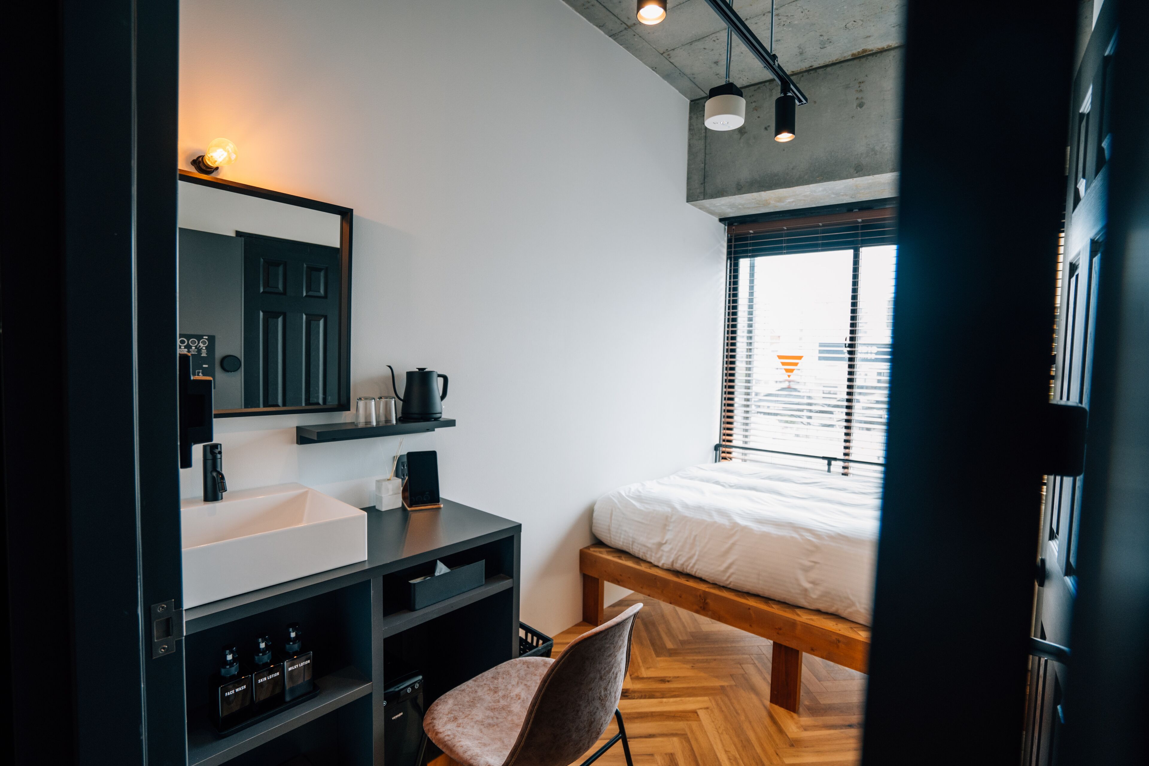Une chambre moderne avec un lit en bois, une table de chevet blanche, un bureau noir avec une chaise, un grand miroir et une fenêtre avec des stores.